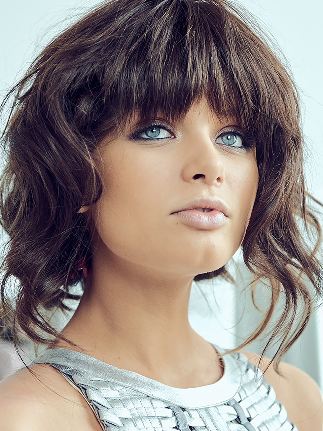 Close-up beauty portrait of a model with striking blue eyes, textured brunette bangs, and a metallic silver top, captured in soft natural light