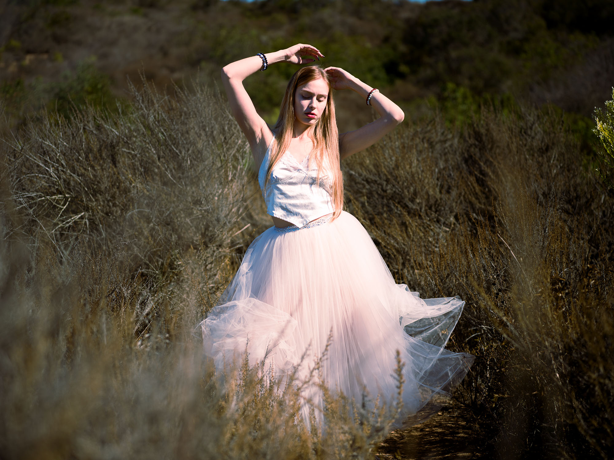 Editorial fashion portrait of a blonde model in a flowing tulle skirt and lace top, posing gracefully in a sunlit natural field setting