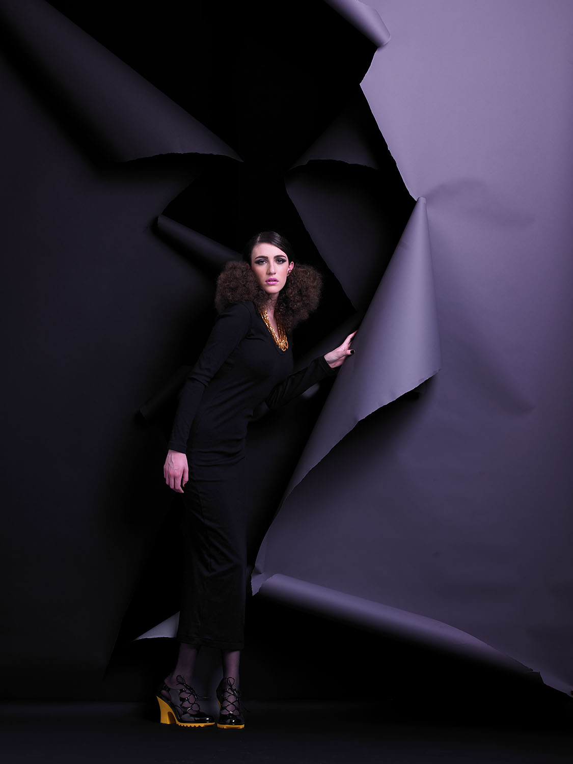High-fashion editorial portrait of Renee Vermeuel in a long black dress and bold yellow heels, standing against a torn paper backdrop in a dramatic studio setting