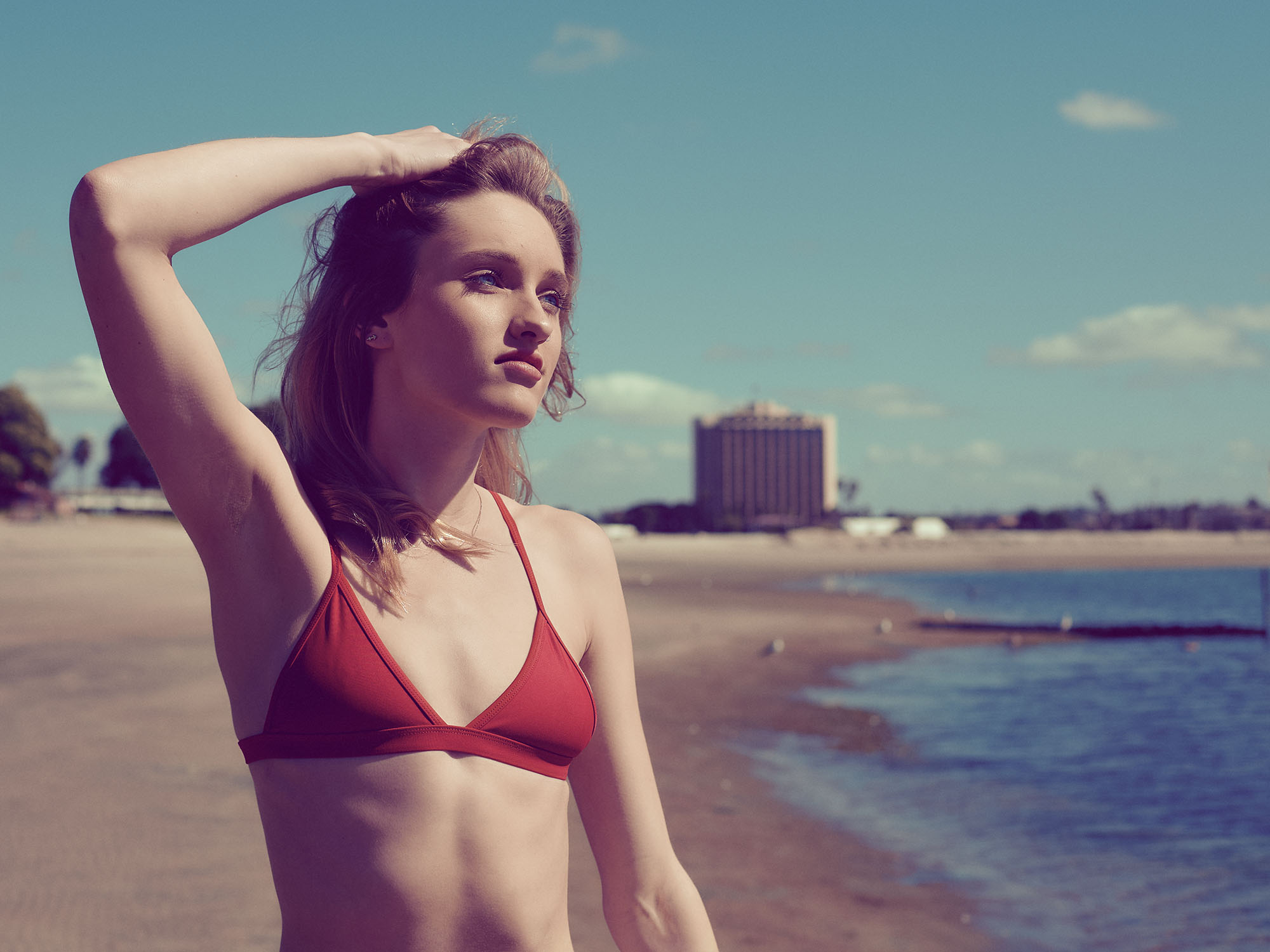 Blonde model in a red bikini posing on the beach under warm afternoon light, with a distant hotel in the background and a cinematic summer mood