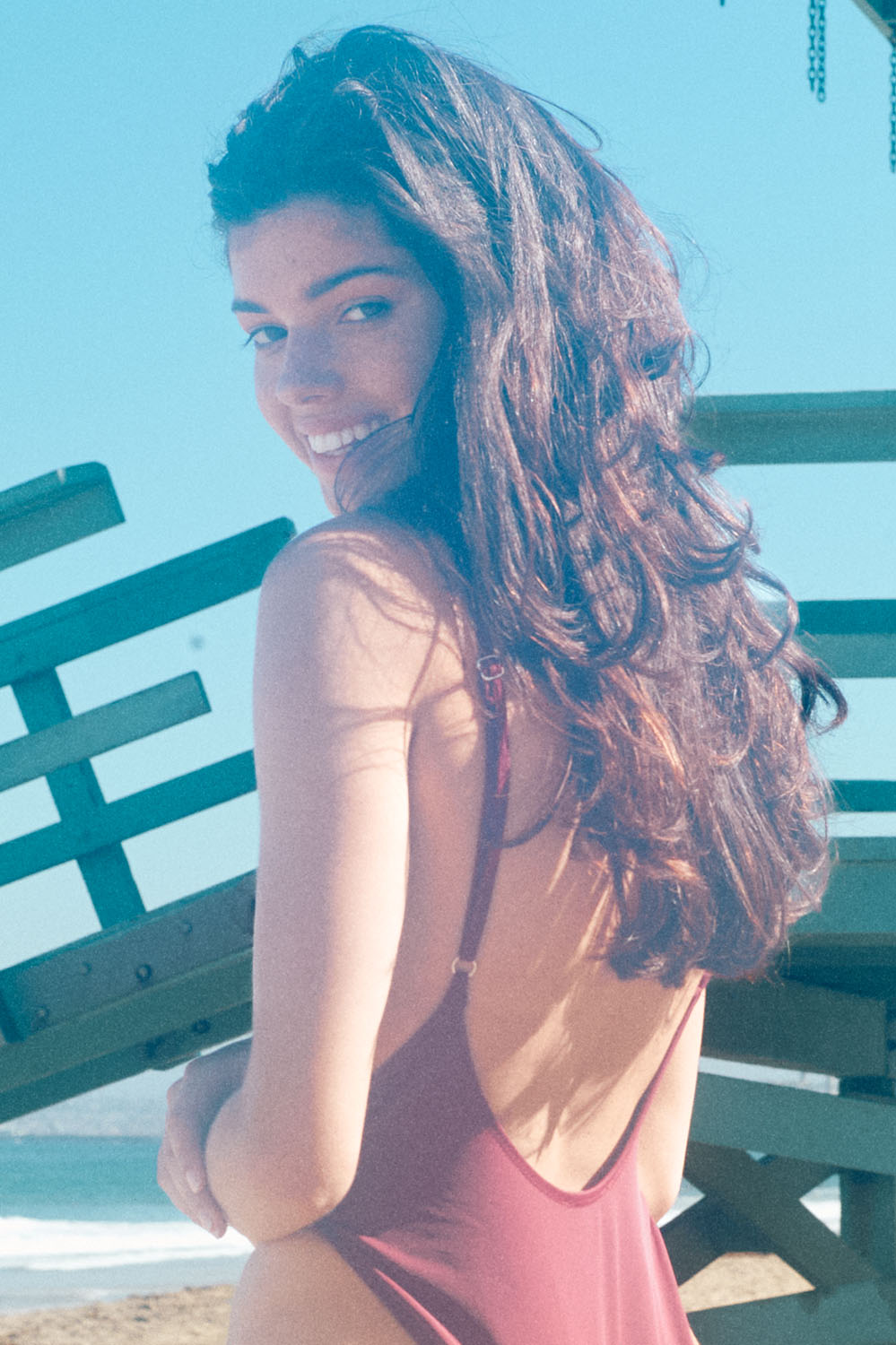 Playful beach portrait of model Mira Chatterjee in a swimsuit, smiling over her shoulder near a lifeguard stand in bright sunlight with a film-style aesthetic