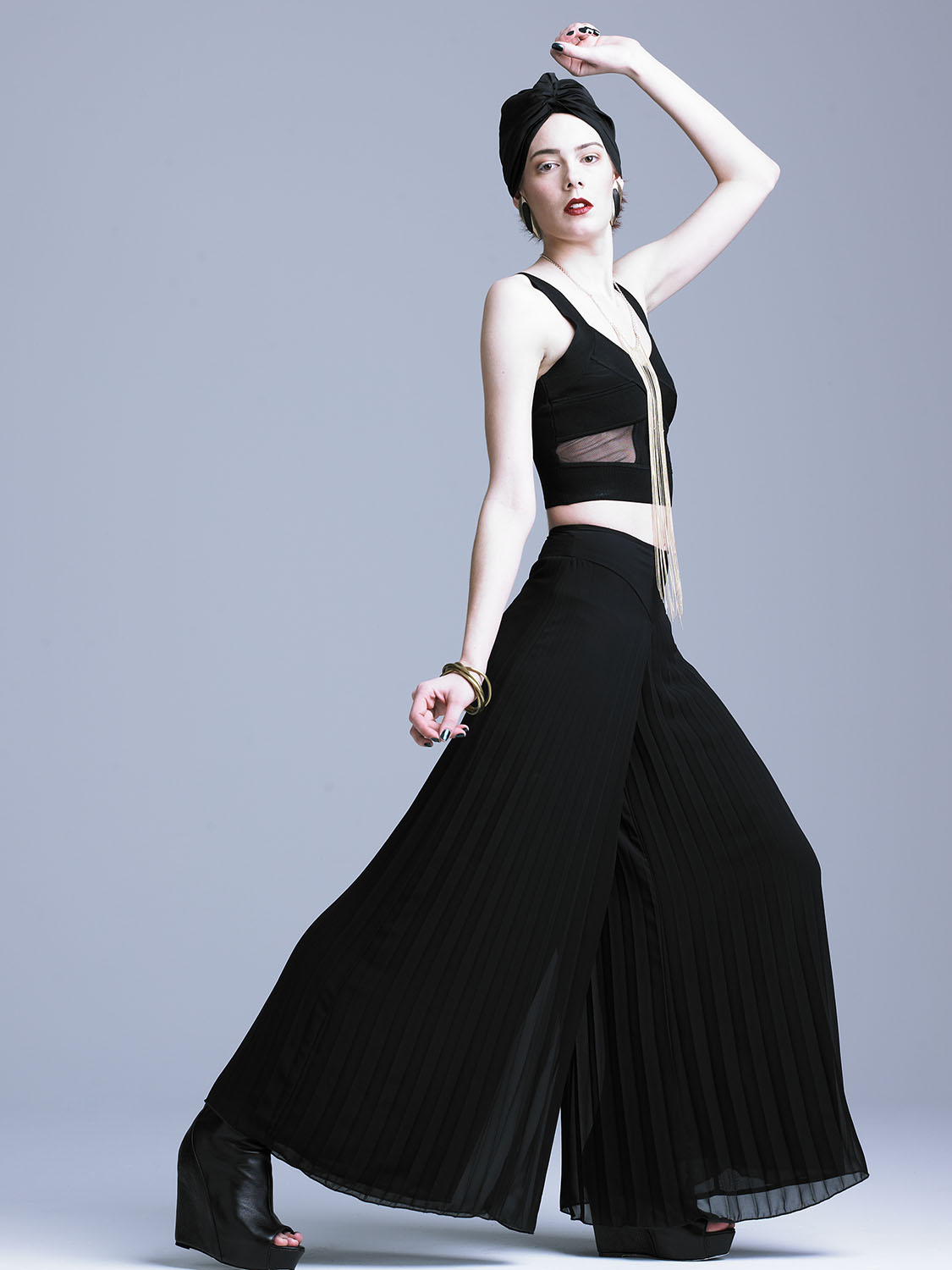 Studio fashion editorial image of a model in a black mesh crop top and pleated palazzo pants, accessorized with gold jewelry and a turban, striking a bold pose