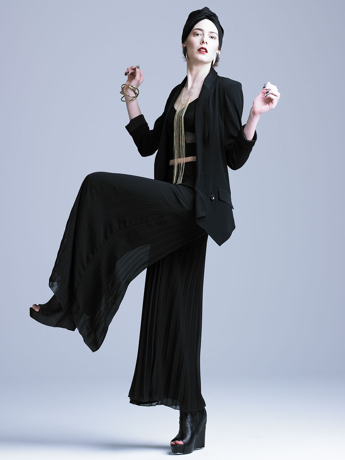 High-fashion studio portrait of a model in a dramatic black outfit with pleated wide-leg pants, gold accessories, and a black turban, captured mid-pose