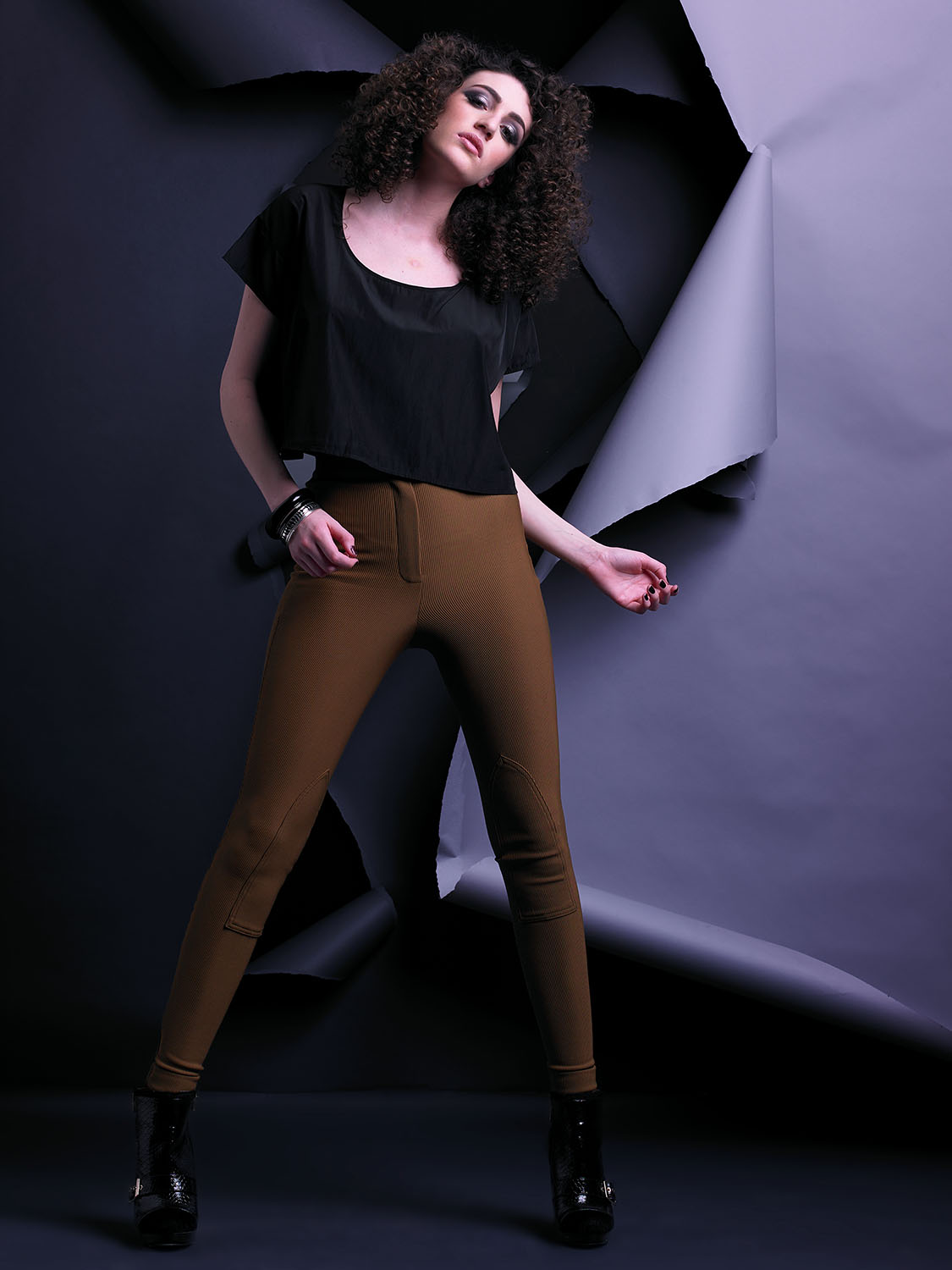 Editorial fashion portrait of Renee Vermeuel with voluminous curly hair, wearing a black top and brown fitted pants against a sculptural dark paper backdrop